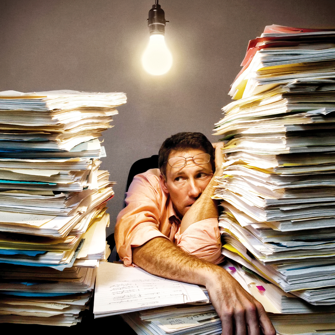 Homme épuisé entouré de piles de papiers sous une ampoule, symbolisant le stress administratif rencontré par les voyageurs ou WHV en Australie